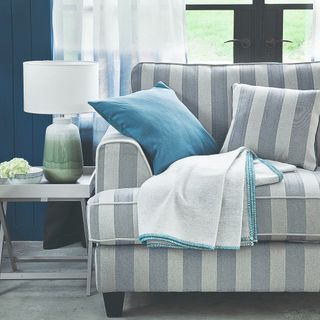 A blue-painted living room with a striped blue and white love seat next to a window with cushions and a throw on top