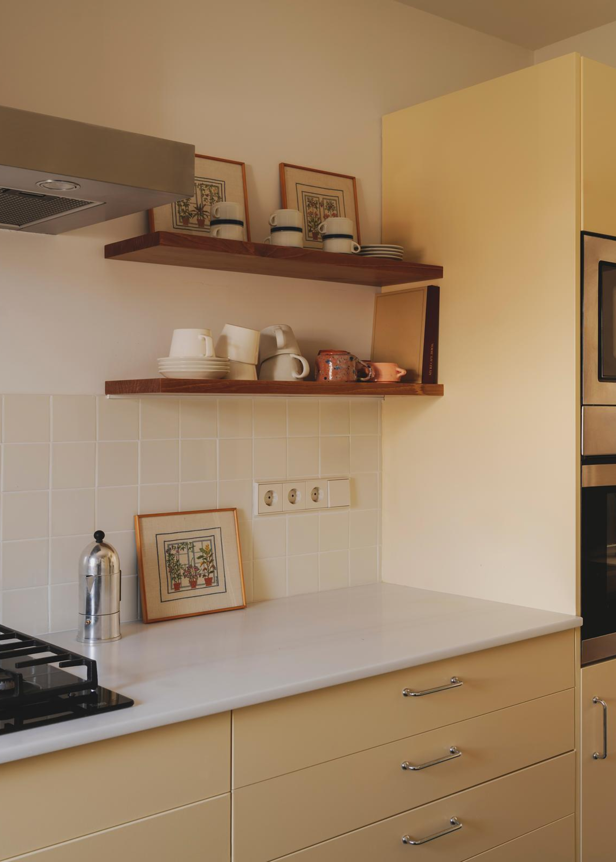 A cozy kitchen with yellow cabinetry , wooden wall-mounted shelving and a white tile backsplash