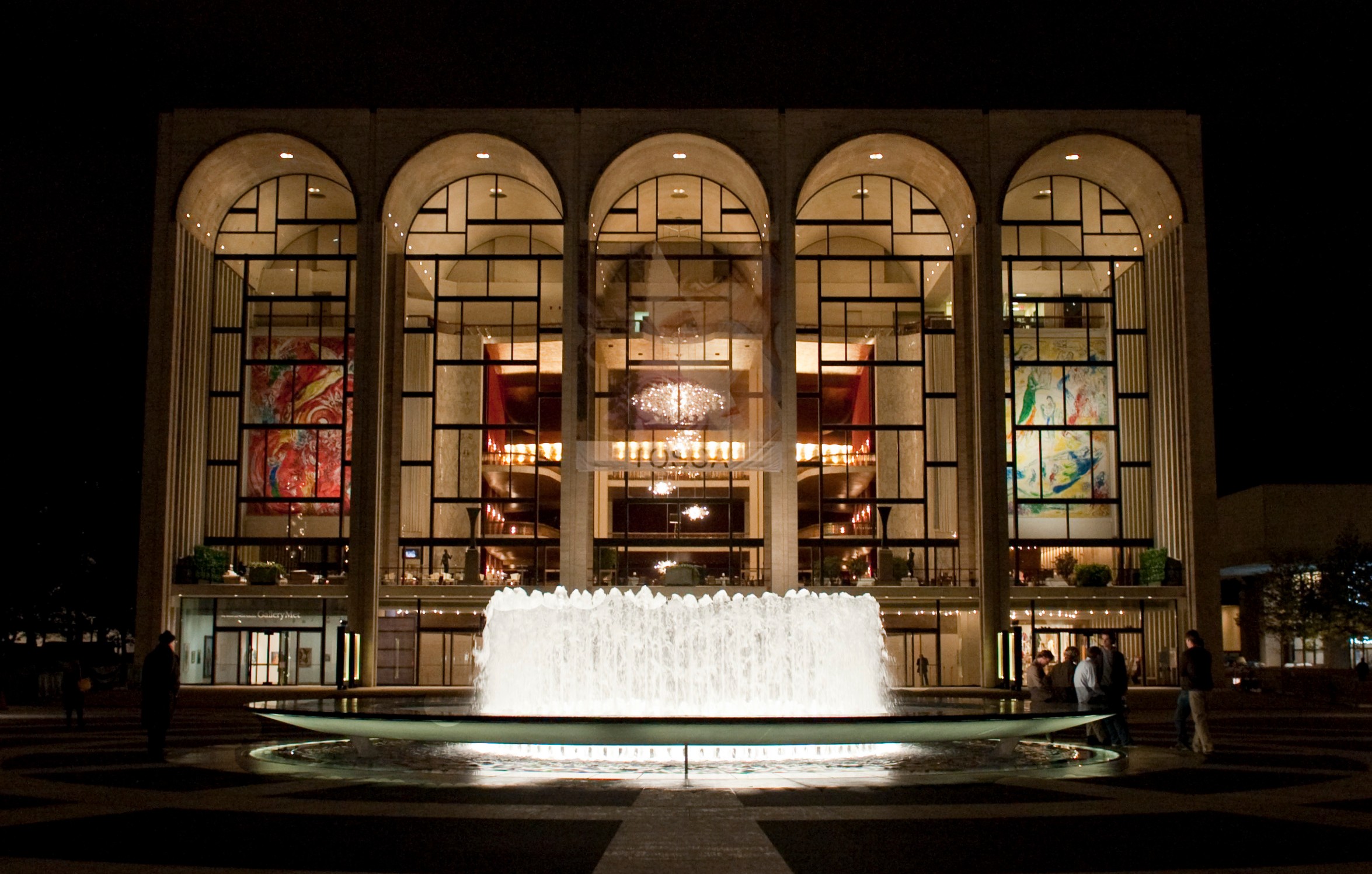 The Metropolitan Opera in New York City lit up at night, as seen from Lincoln Center Plaza with a fountain in front of it. 