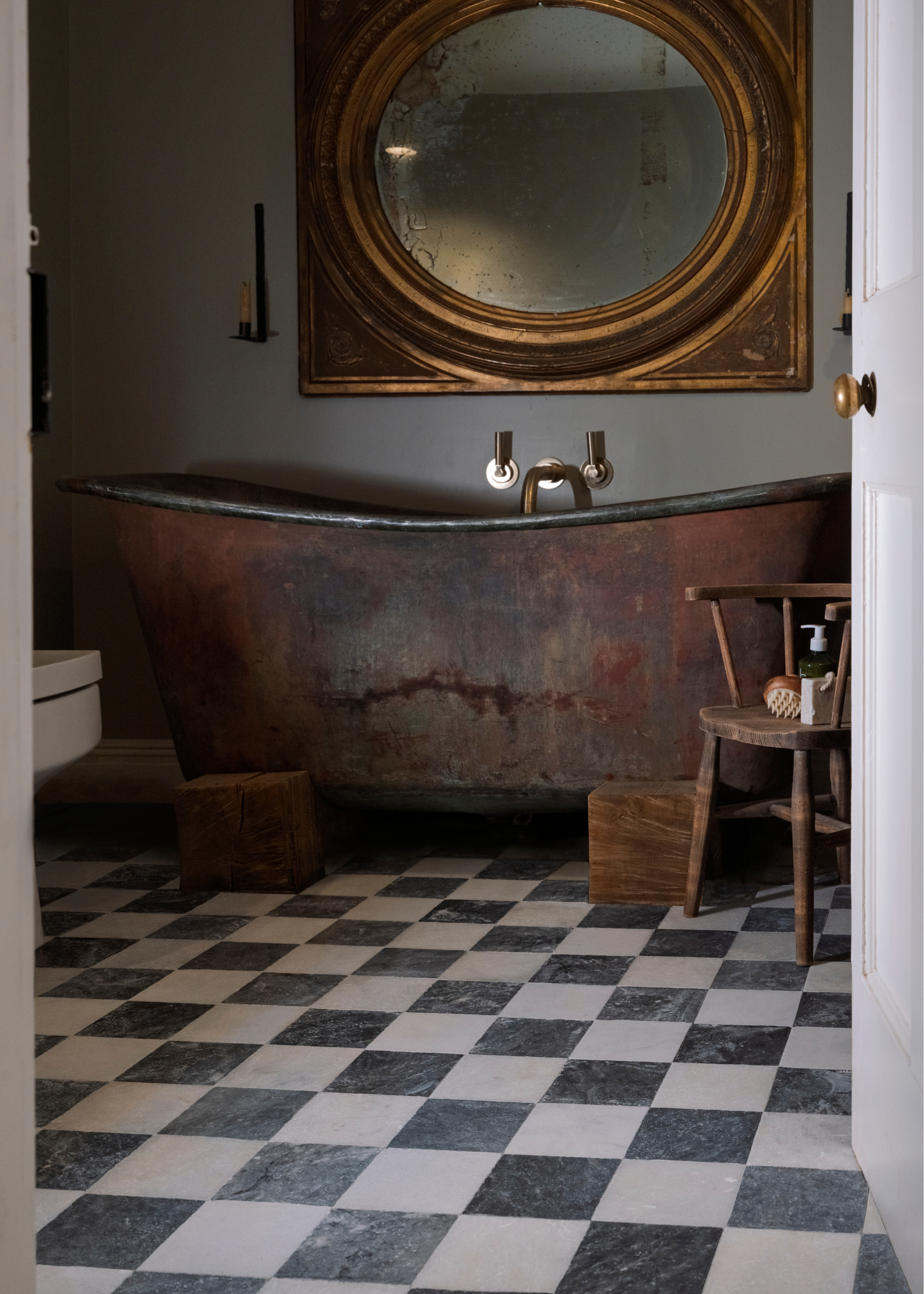 A small bathroom full of raw rugged materials that look like antiques including a copper bath tub on wooden sleepers, a large brass mirror above the bath and an old wooden chair wuth toiletries on, all sitting on a chequerboard tile floor