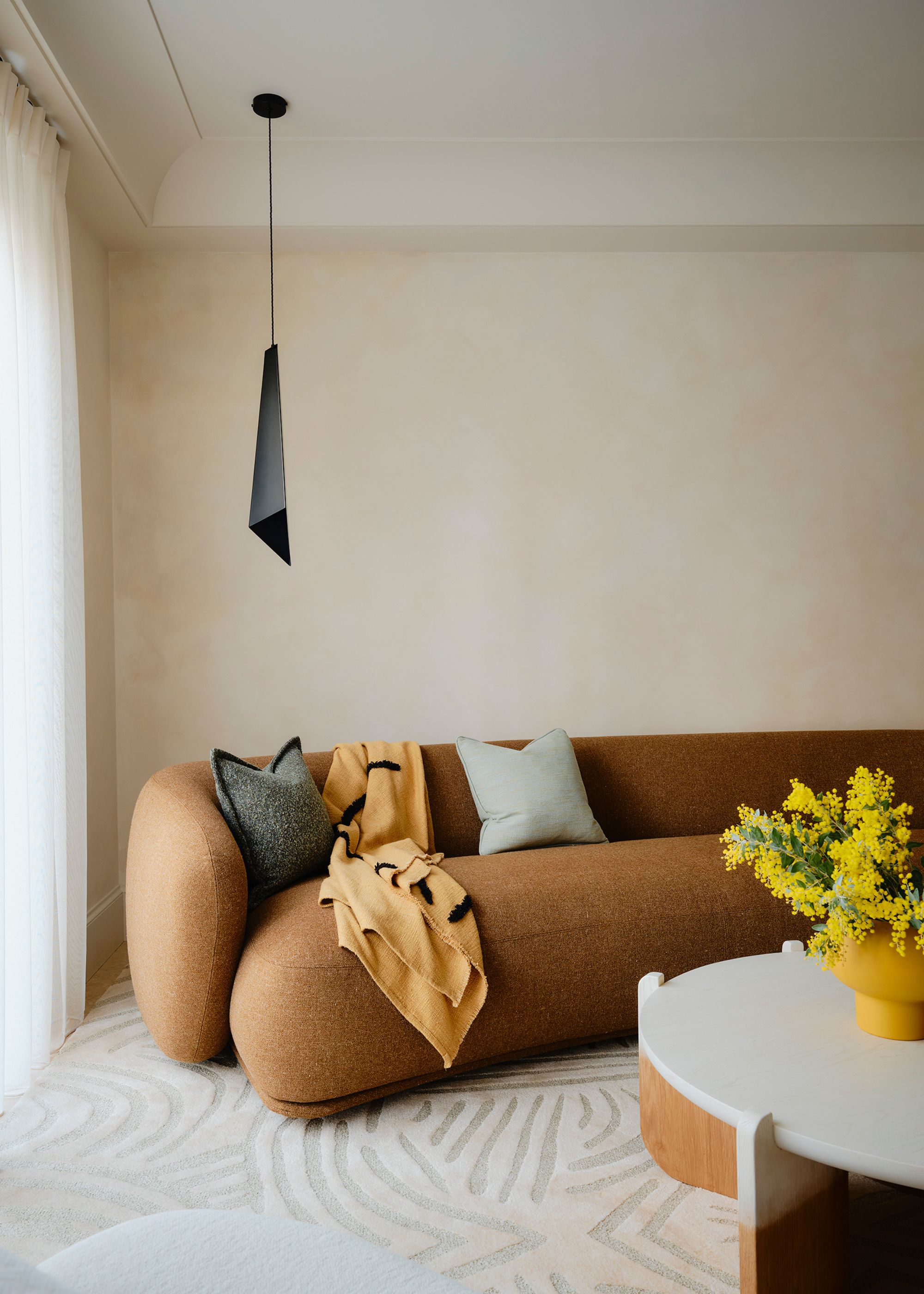 A living room with warm white limewash walls, a modern sculptural brown sofa, a cream rug, and a coffee table with a vase of yellow flowers.