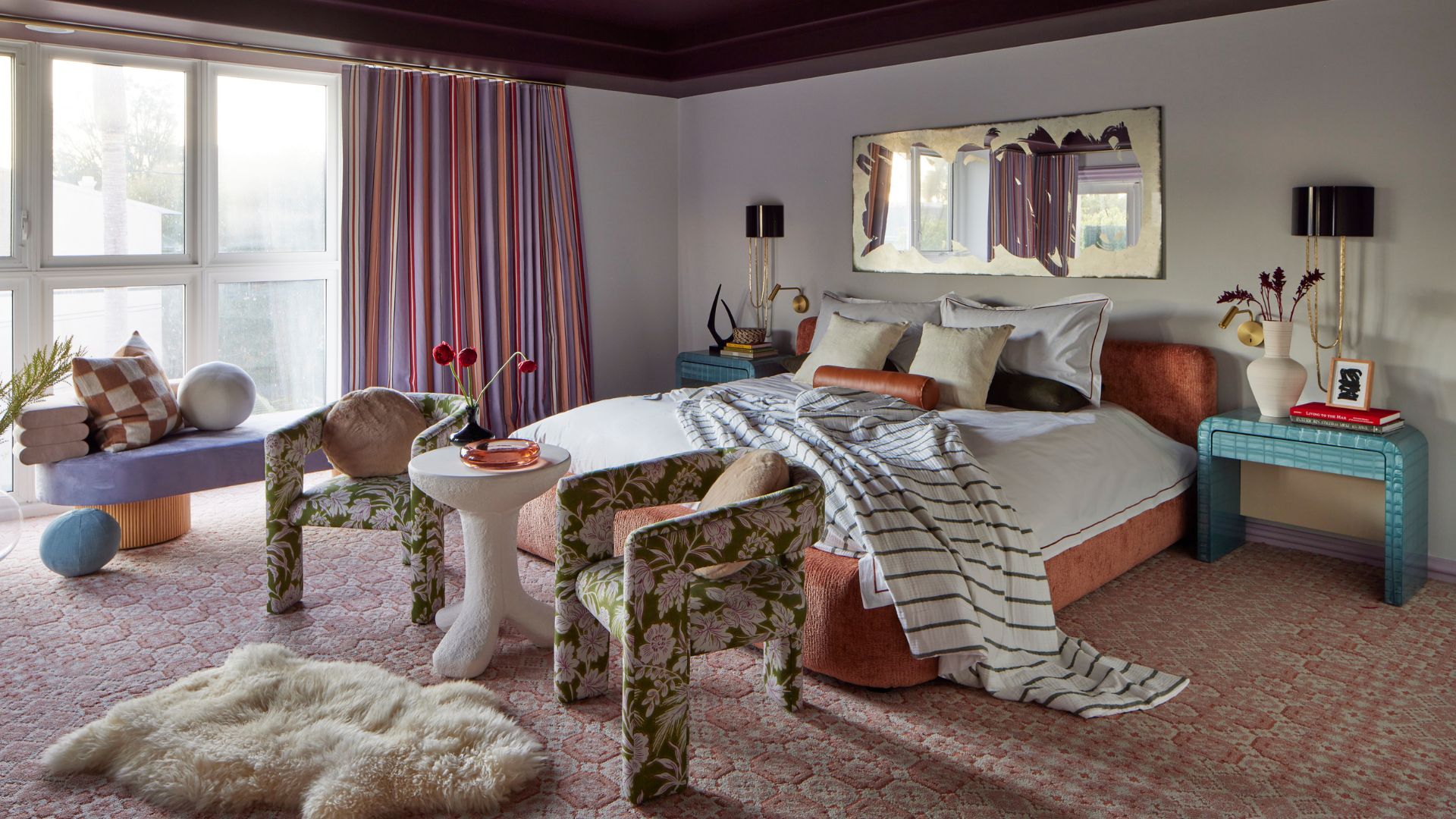 A colorful bedroom with a large bed, tiled side tables, a pair of patterned chairs with a white table in the middle, a faux fur floor rug, a bench with cushions and a statement mirror