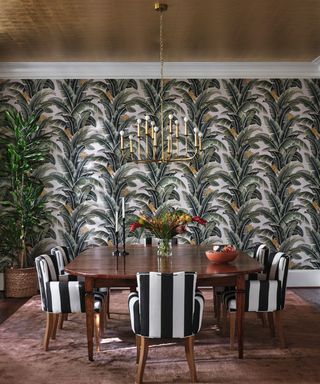 A dining room with leafy wallpaper, a wooden dining table, and black-and-white striped, upholstered armchairs