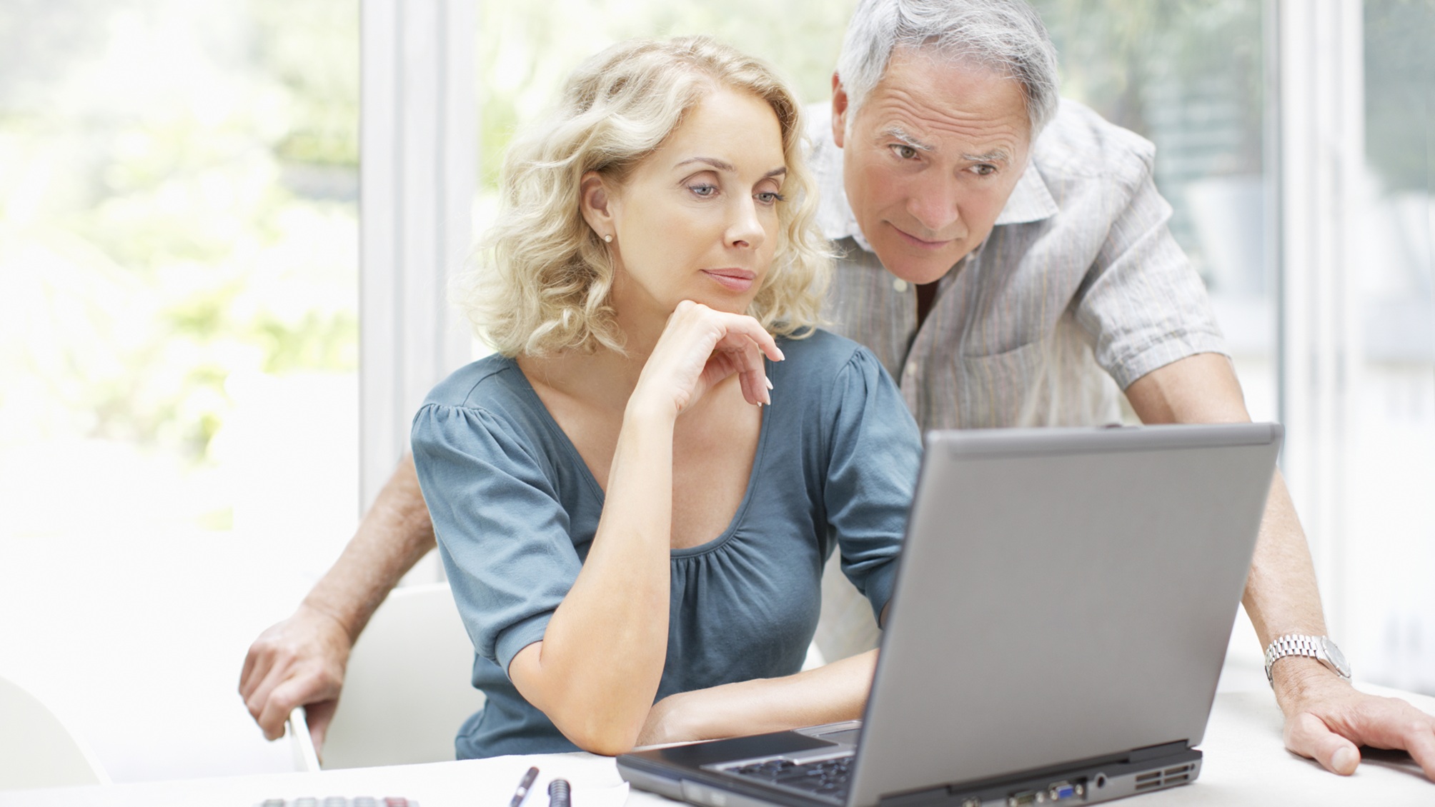 An older couple look at a laptop together in their kitchen.