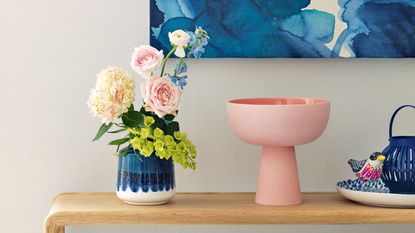 Light wood bench against an off-white wall with a blue vase holding pink flowers, a sculptural pink vase and a blue abstract canvas on the wall