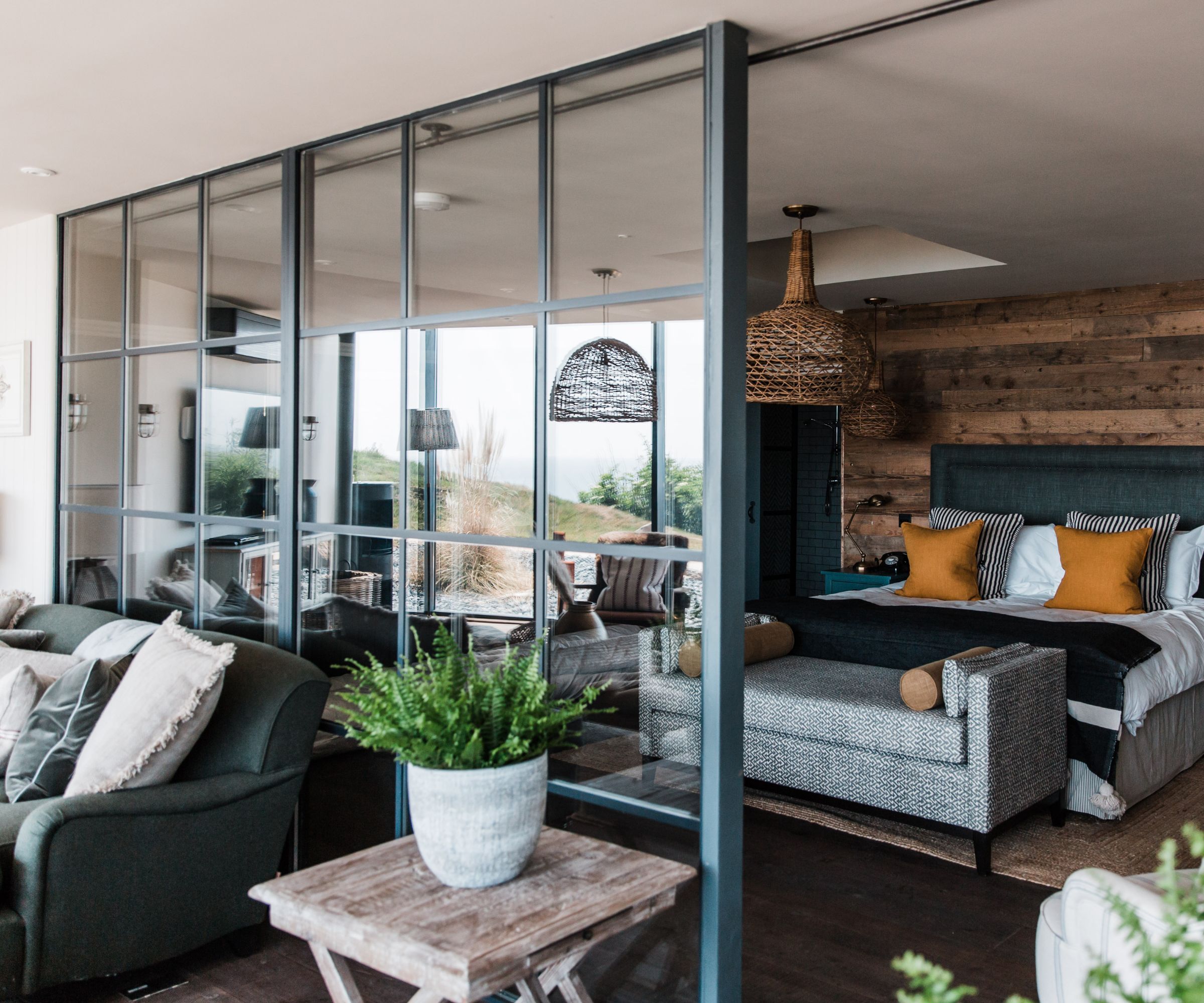 A stylish open-plan bedroom divided by a black metal-framed glass partition. On one side sits a cozy living area with dark armchairs and soft cushions. On the other, a bed with mustard and striped pillows rests against a rustic wood-paneled wall, beneath woven pendant lights.