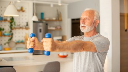 waist-shot of a man standing looking off camera holding two small blue dumbbells with arms outstretched in front. there's a kitchen setting behind him.