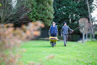 Two men walking across a lawn, one with a wheelbarrow, as they garden during cold winter months