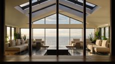 solarium in house with skylights, glossy tiled floor, beige sofas and a coffee table in the middle, floor-to-ceiling windows that look out to the lake