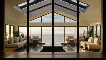 solarium in house with skylights, glossy tiled floor, beige sofas and a coffee table in the middle, floor-to-ceiling windows that look out to the lake
