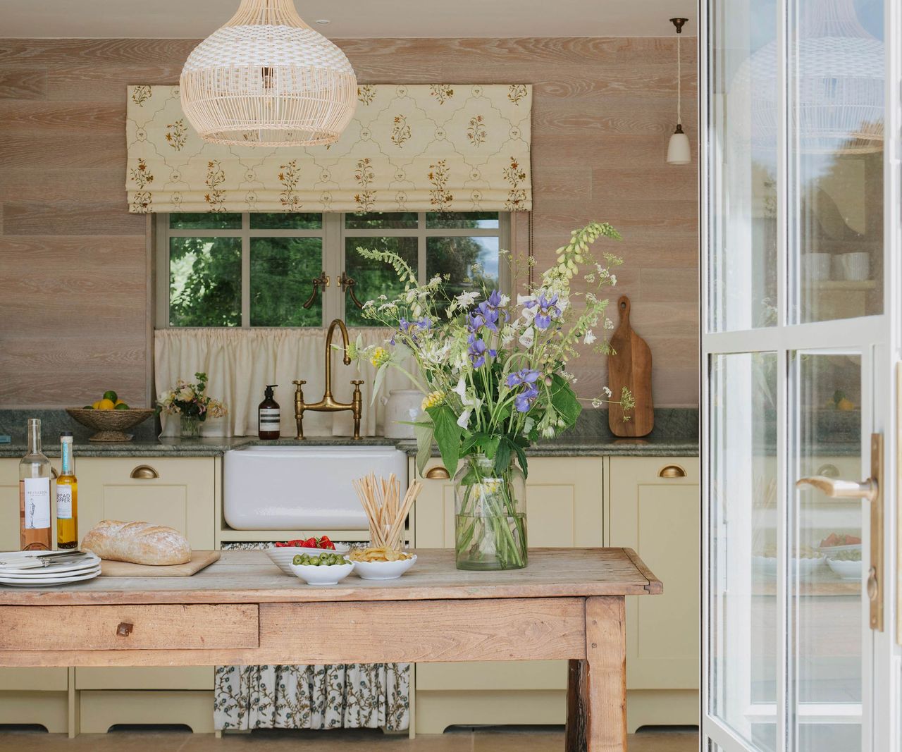 A pool house kitchen with pale yellow cabinetry, a Belfast sink with brass bridge tap, floral Roman blind, rattan pendant light and a rustic wooden table with wildflowers.