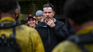 Kevin Alejandro holding up his right hand while directing two people in firefighter outfits, Stephanie Arcila is standing behind him smiling.