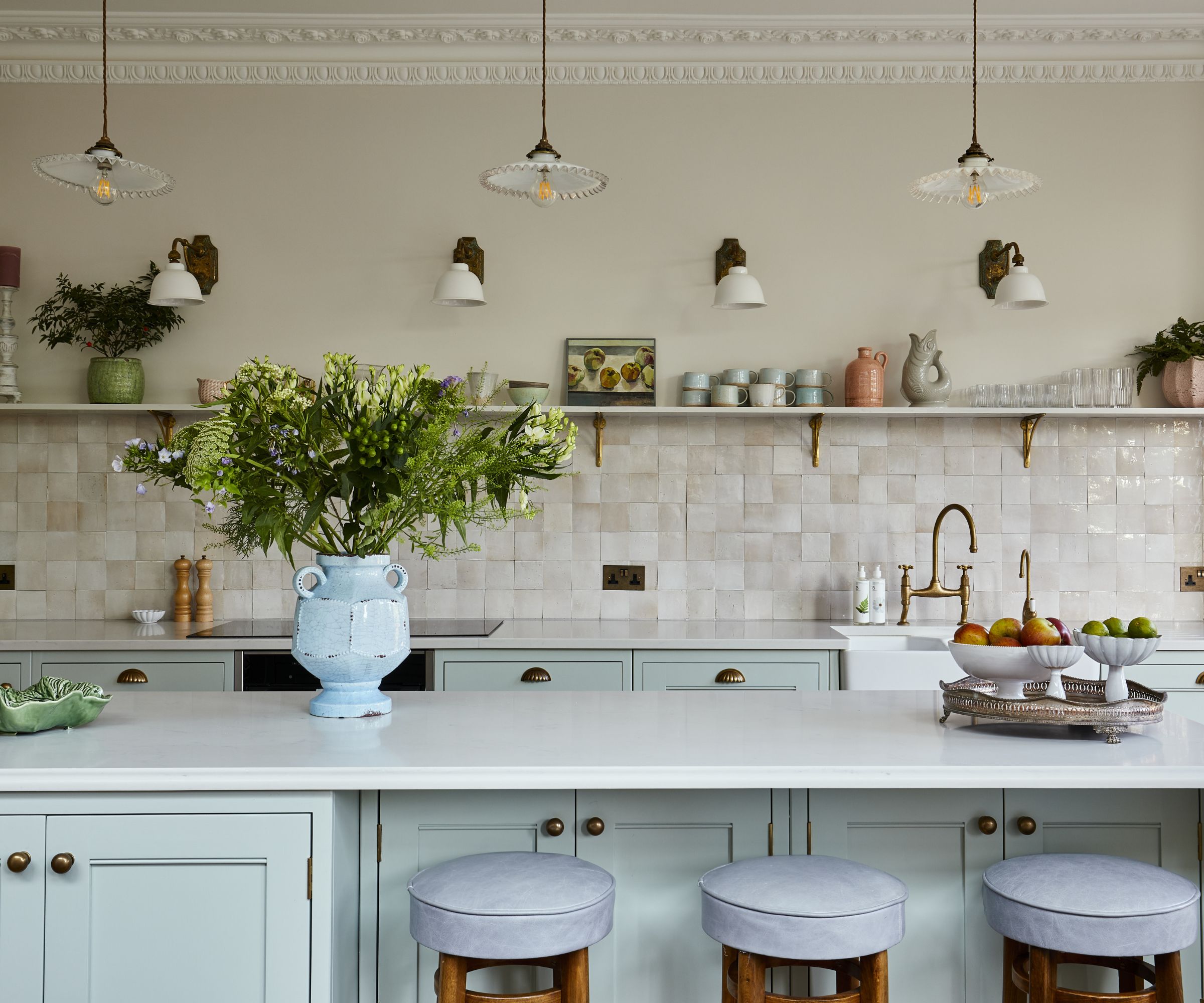 Kitchen with pale blue cabinetry, a kitchen island with a white countertop, a cream zellige tiled backsplash with a long shelf running along the top decorated with ceramics and three pleated pendants