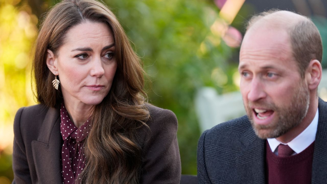 Kate Middleton wears a brown blazer with a burgundy and white polka dot blouse, and has long wavy brown hair, as she looks at Prince William, who is wearing a suit and tie and has beard stubble