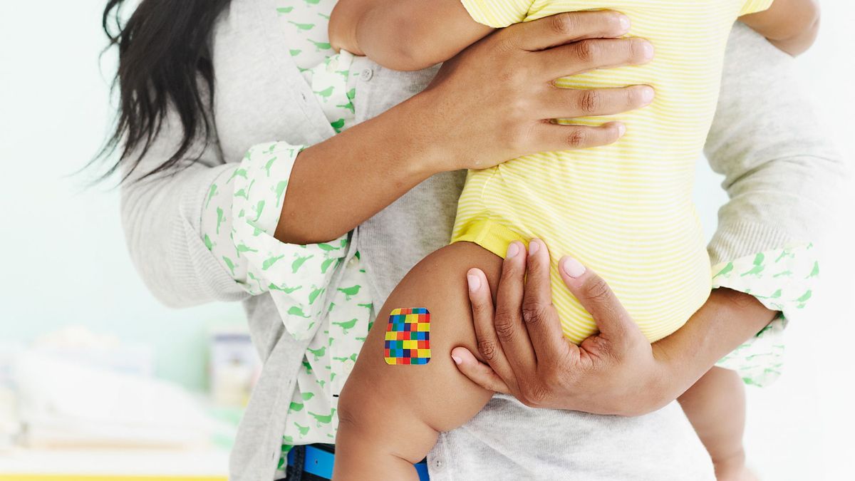 photo shows a woman&#039;s arms holding a baby in a yellow onesie, who has an adhesive bandage on her outer thigh as if the baby just got an injection 