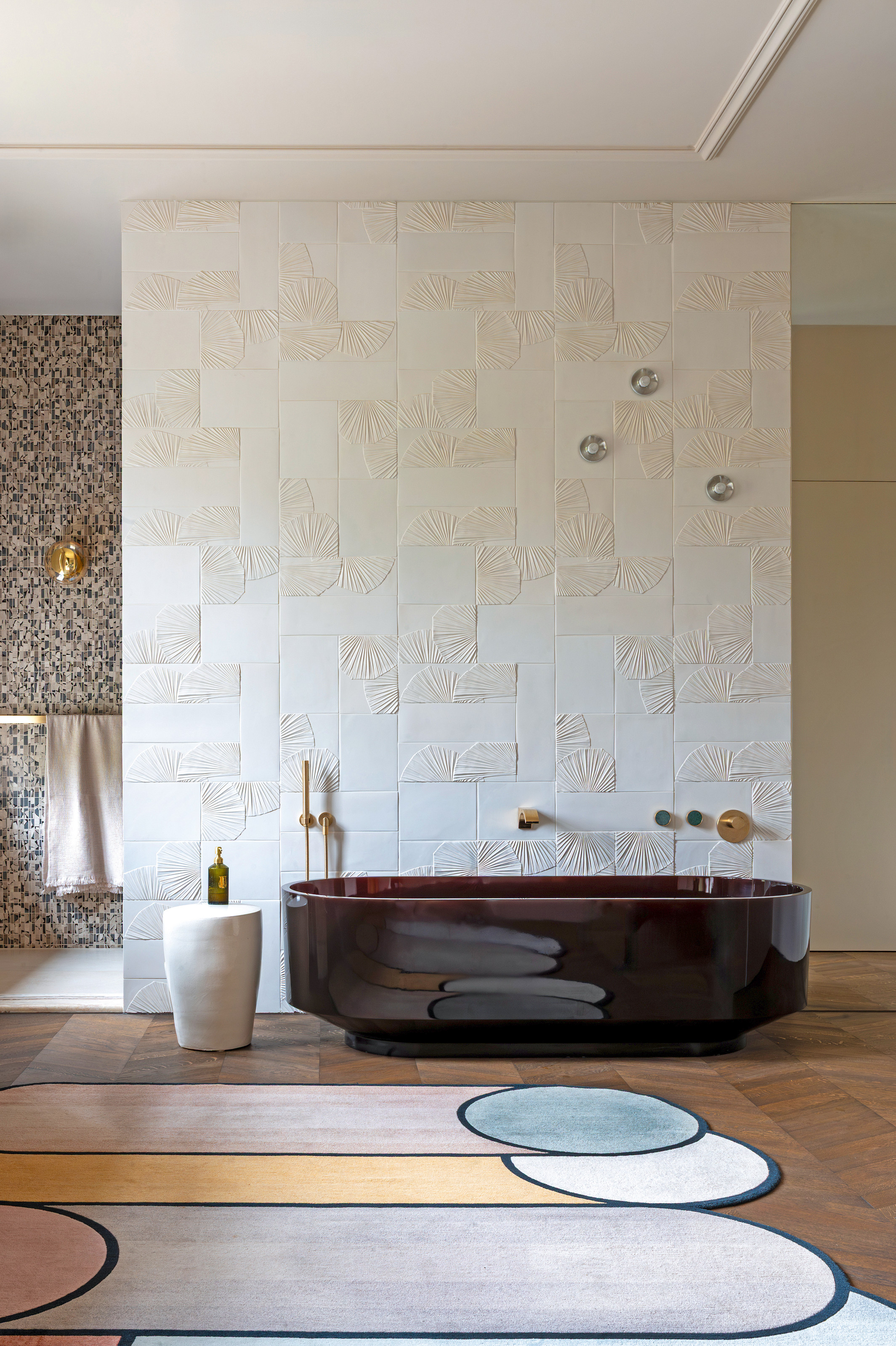 White bathroom with textured wall tiles, parquet wood floor, brown glass bath, abstract rug and white stool
