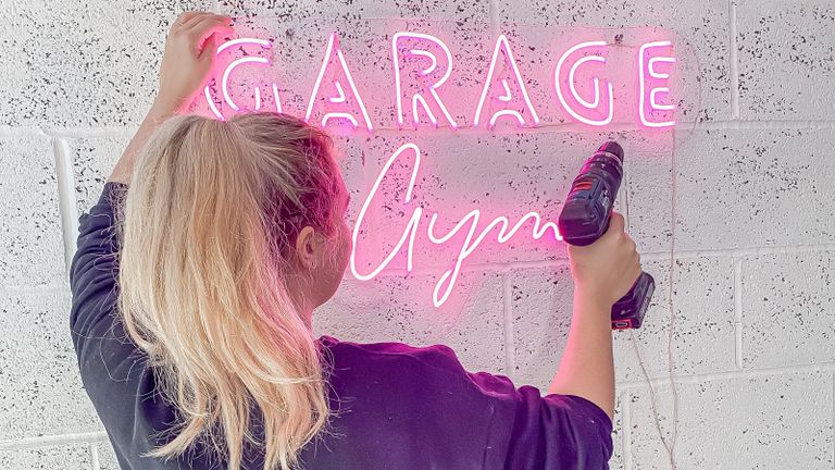 An image of Jasmine Gurney holding up pink DIY LED neon sign which says 'Garage gym' holding electric drill