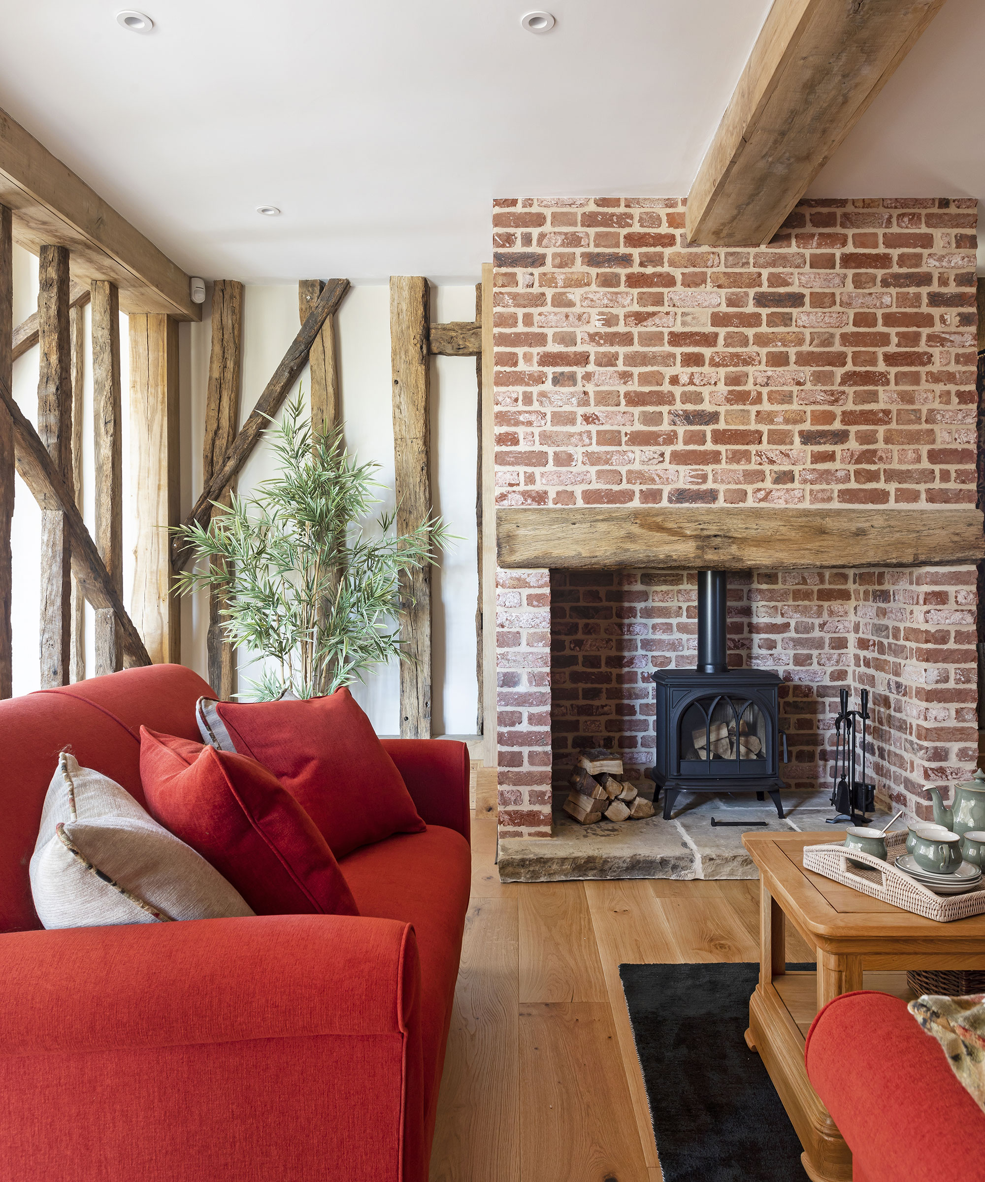 living room with red sofas, wooden floors, oak ceiling beams and a brick inglenook fireplace with a log burner