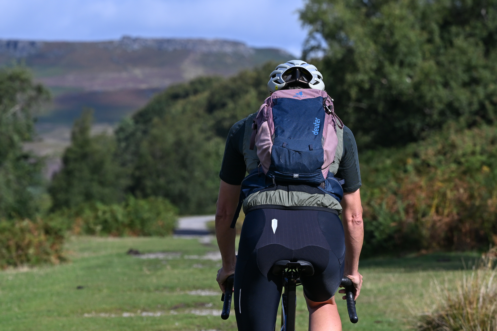 Man riding a gravel bike wearing a green jersey, black shorts and a green gilet with a pink and blue backpack on, riding away from the camera