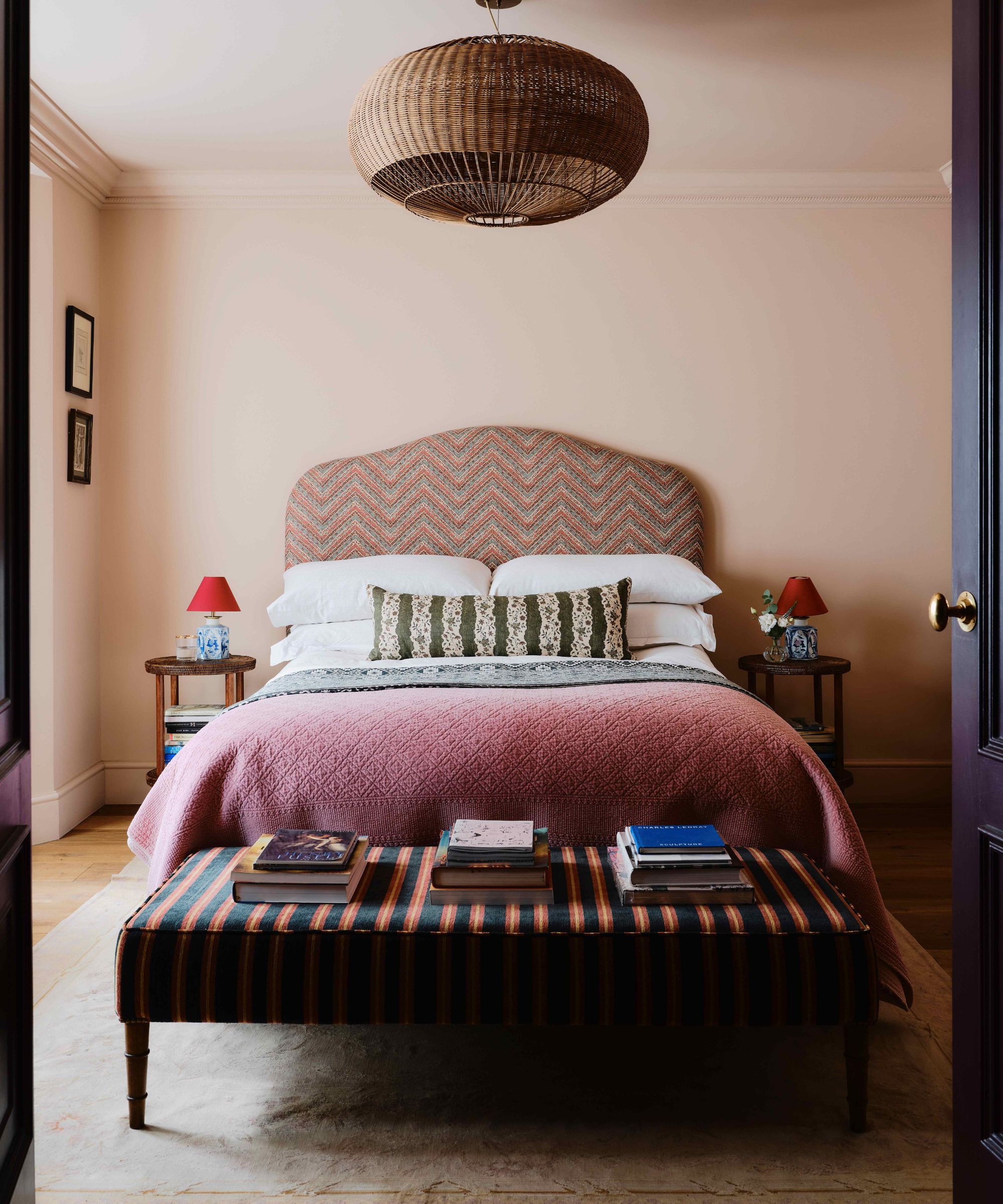 light pink bedroom with a dusty pink patterned headboard with floral lumbar pillow, pink throw blanket, striped ottoman at the foot of the bed and rattan ceiling light