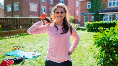 Smiling woman stands in sunny garden holding kettlebell by one shoulder