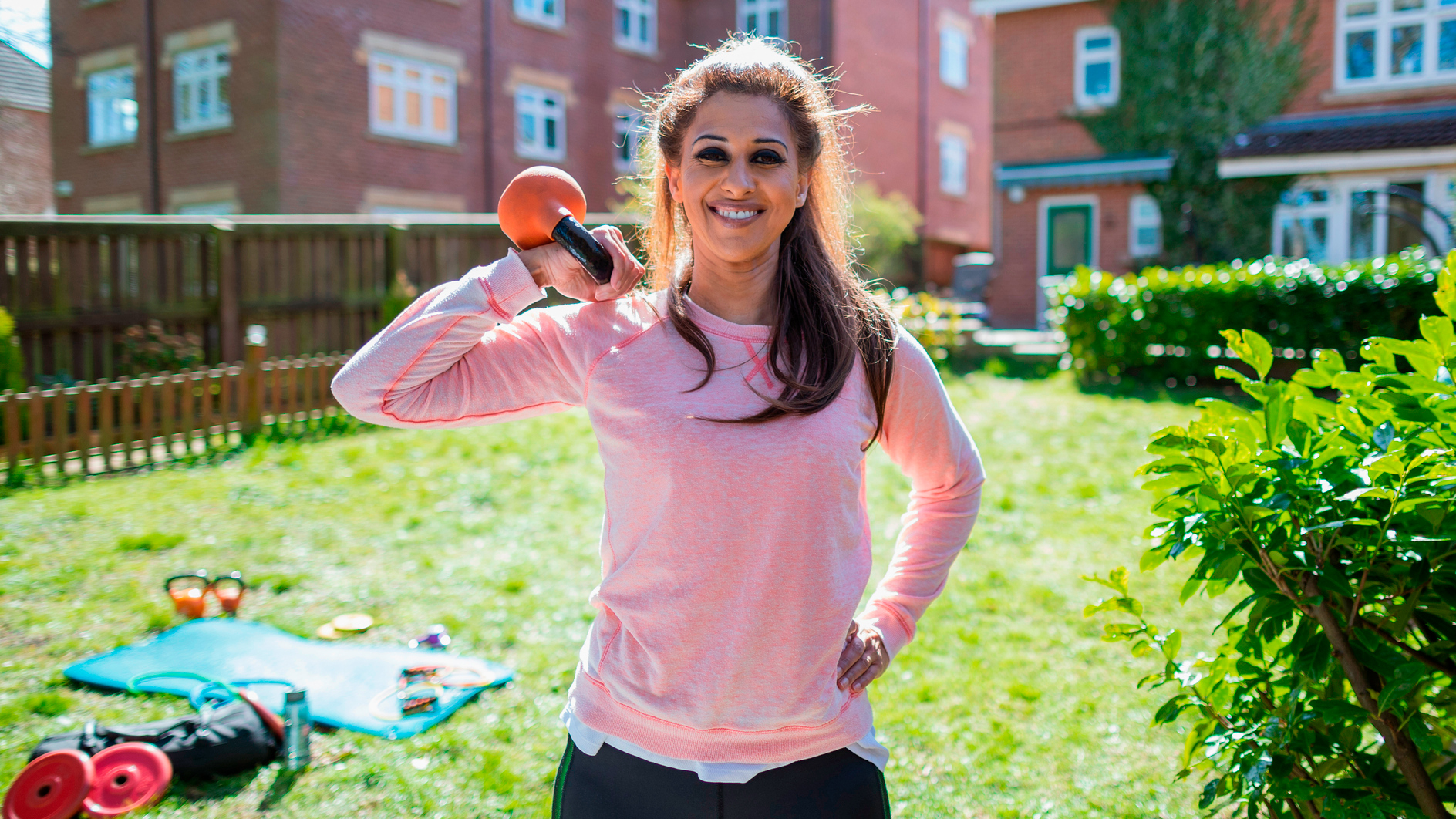 Smiling woman stands in sunny garden holding kettlebell by one shoulder