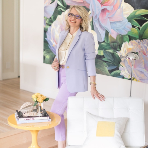 Image of a woman wearing a lavender blazer and pants with a yellow blouse. She is standing by a white accent chair in front of a large painting of flowers.