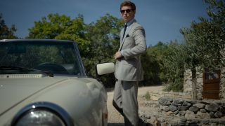 A sunglassed Eddie Redmayne stands in a grey suit next to his sports car in The Day of the Jackal.