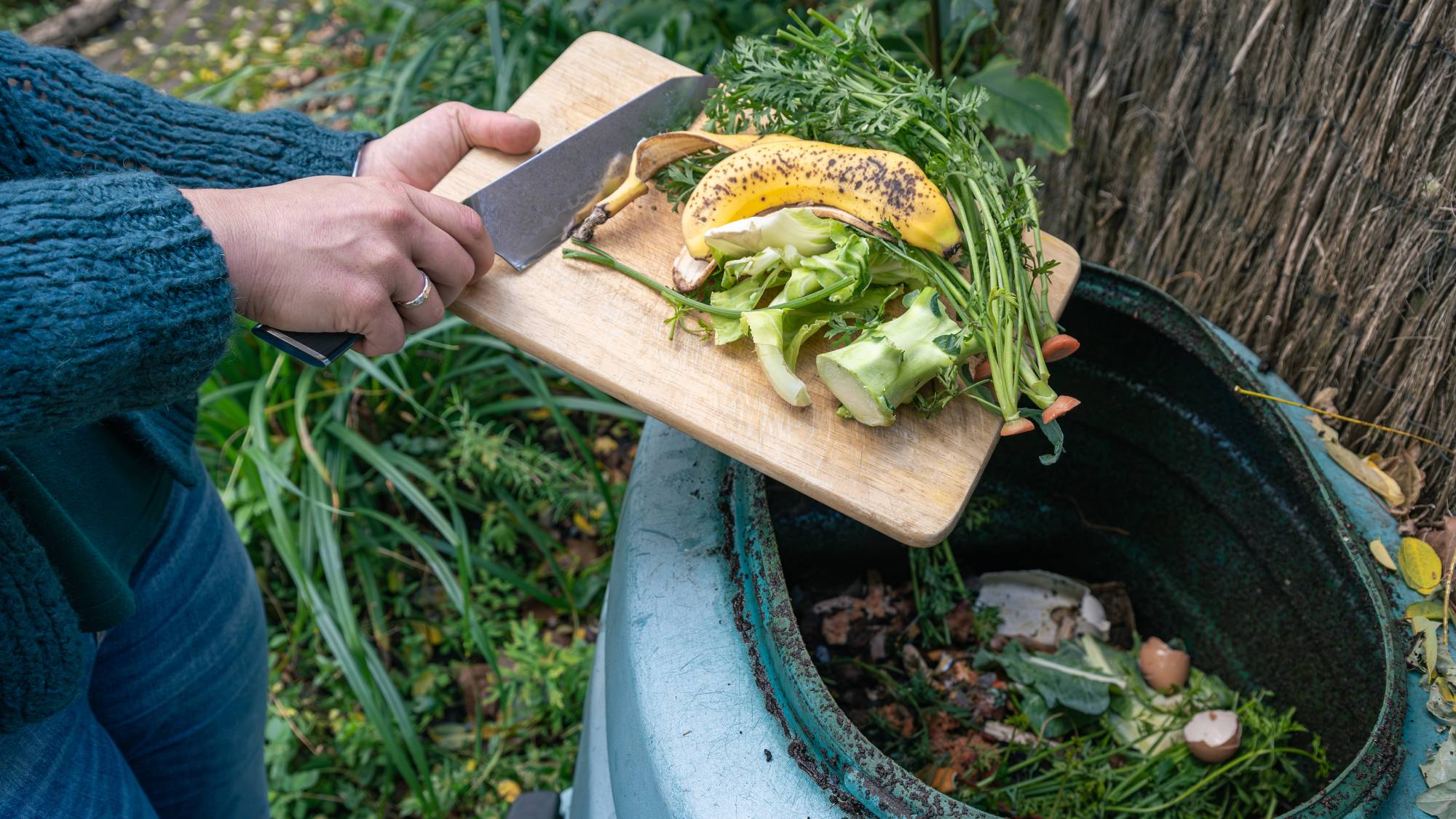 Hands using a knife to scrape kitchen scraps off a cutting board into a composter