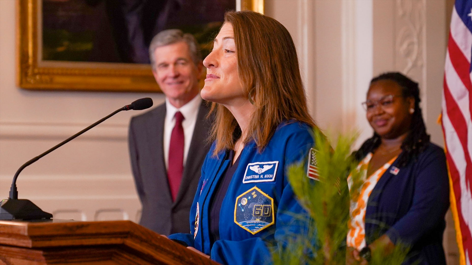 a woman in a blue flight suit stands at a podium with two smiling people and the american flag behind her.