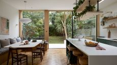 An open kitchen and dining space with floating shelves holding plants over a white island and round stools. Also pictured is a banquette dining space with a white dining table and matching chairs