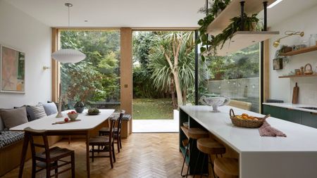 An open kitchen and dining space with floating shelves holding plants over a white island and round stools. Also pictured is a banquette dining space with a white dining table and matching chairs