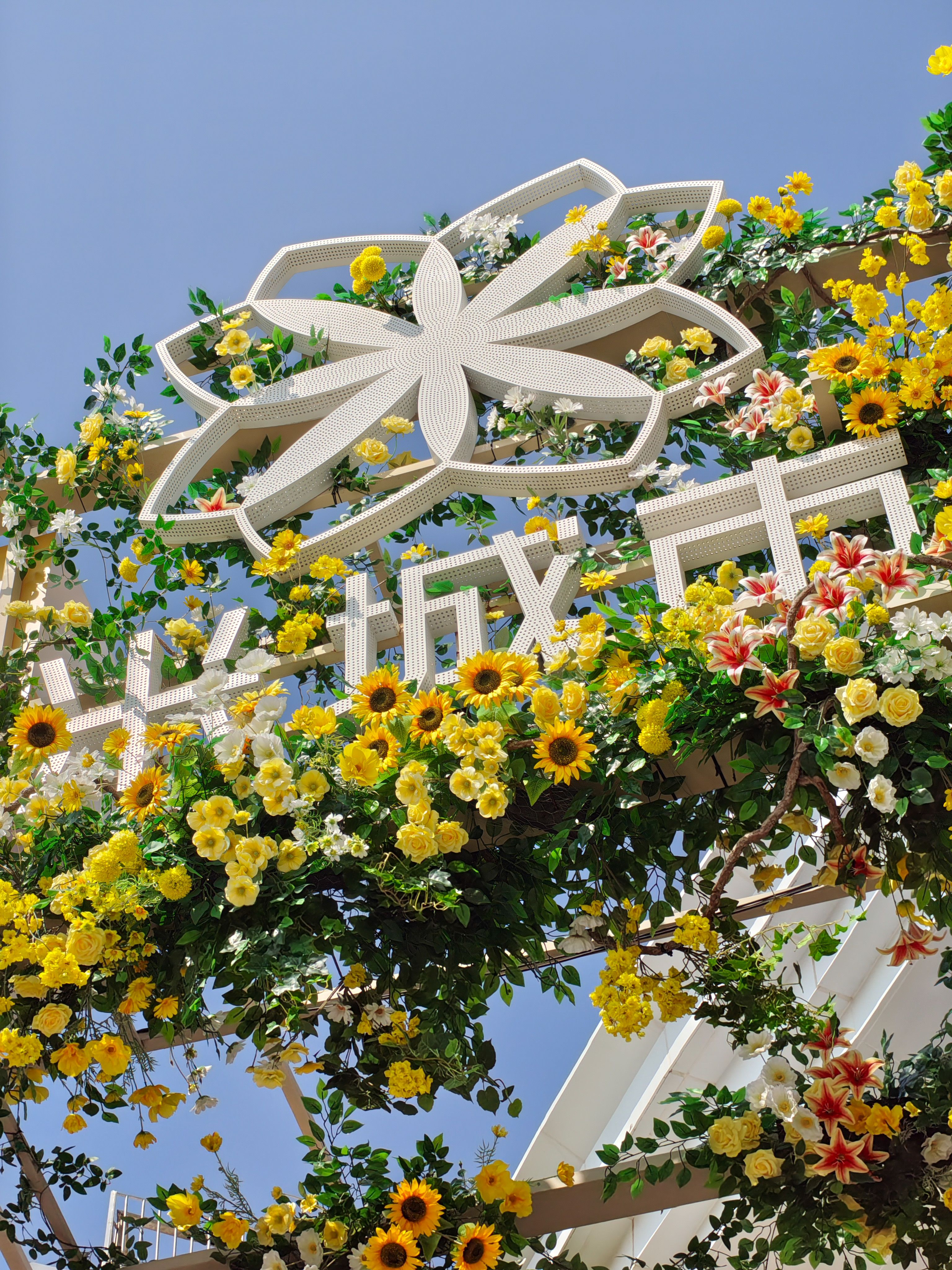 A medium shot looking up at a decorative white architectural structure adorned with many yellow sunflowers and white lilies. The structure features a large, stylized eight-petal flower at the top and Chinese characters below it. The scene is set against a bright, cloudless blue sky, emphasizing the contrast between the white metal and the vibrant yellow flowers.