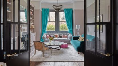 pastel living room ideas, pastel pink living room with turquoise blue drapes and couch, chandelier, grey sofa, perspex table lamp, glass coffee table, pastel and print cushions, grey rug, built in shelving in alcove, view through crittall doors