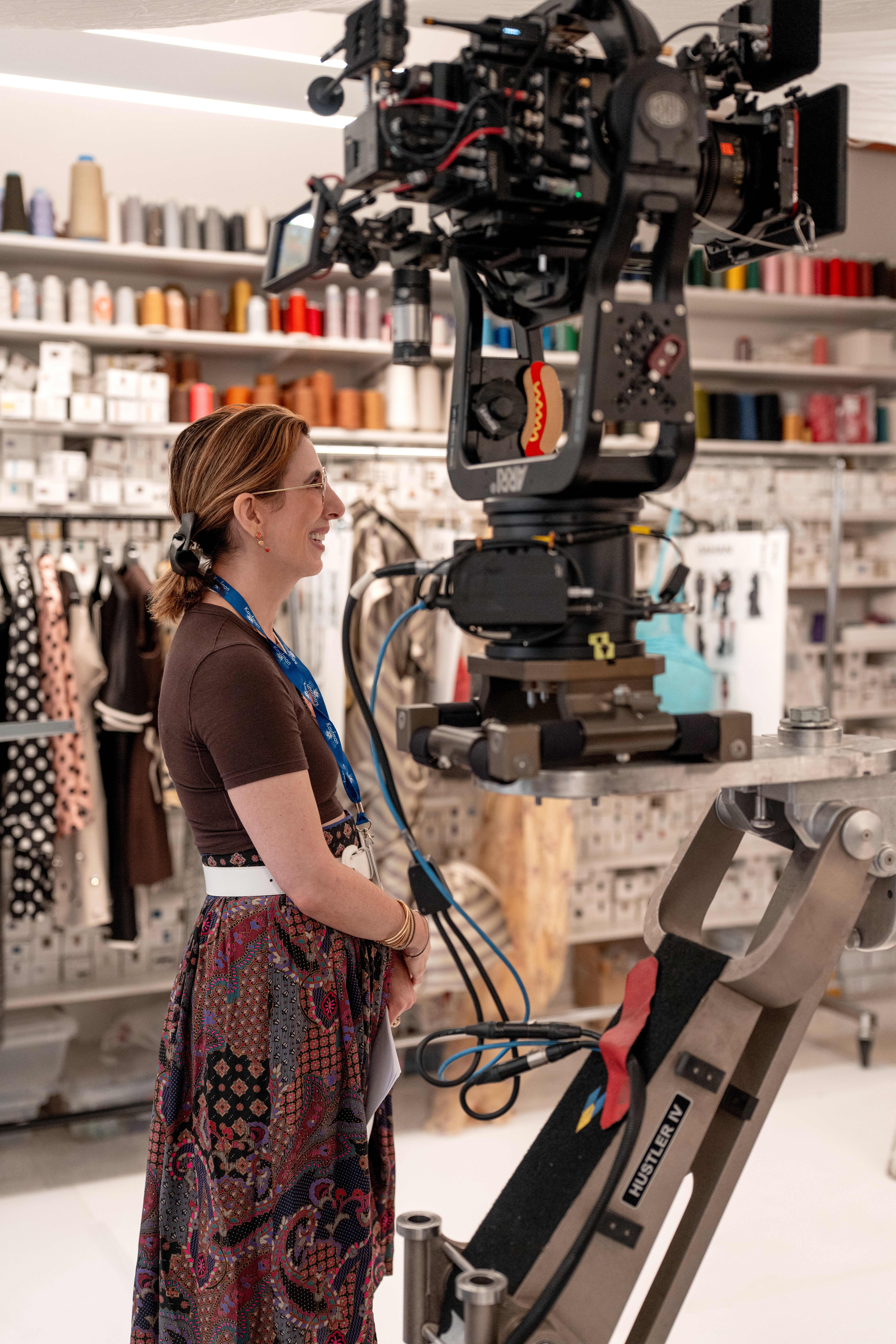 Aline Brosh McKenna wearing a brown tshirt and printed skirt smiling standing by a camera on the set of THE DEVIL WEARS PRADA 2