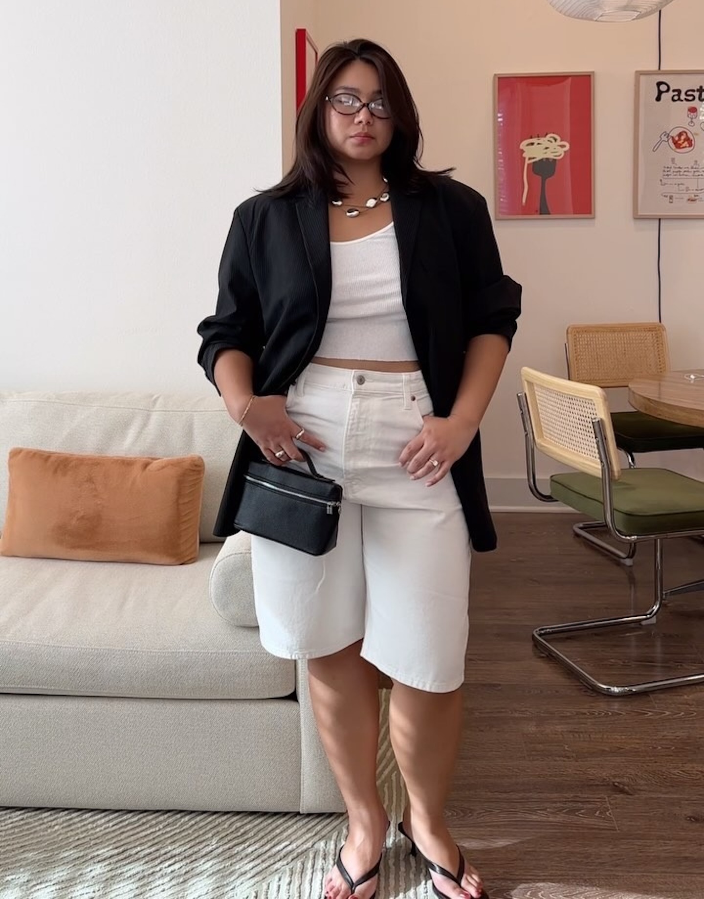 style influencer Marina Torres poses in her living room wearing oval optical eyeglasses, an oversize black blazer, a silver statement necklace, a white tank top, high-waisted white Bermuda denim shorts, mini black bag, and black kitten-heel flip-flops