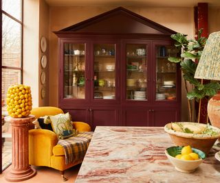 A freestanding kitchen dresser with a cupboard hat detail painted in a rich burgundy hue. The walls of the dining area are painted plaster pink, with a yellow armchair placed in the corner and a green and yellow checkered throw covering the seat cushion. A warm pink tones marble table sits at the center of the room, topped with a terracotta table lamp with a pleated green patterned lampshade and two fruit bowls