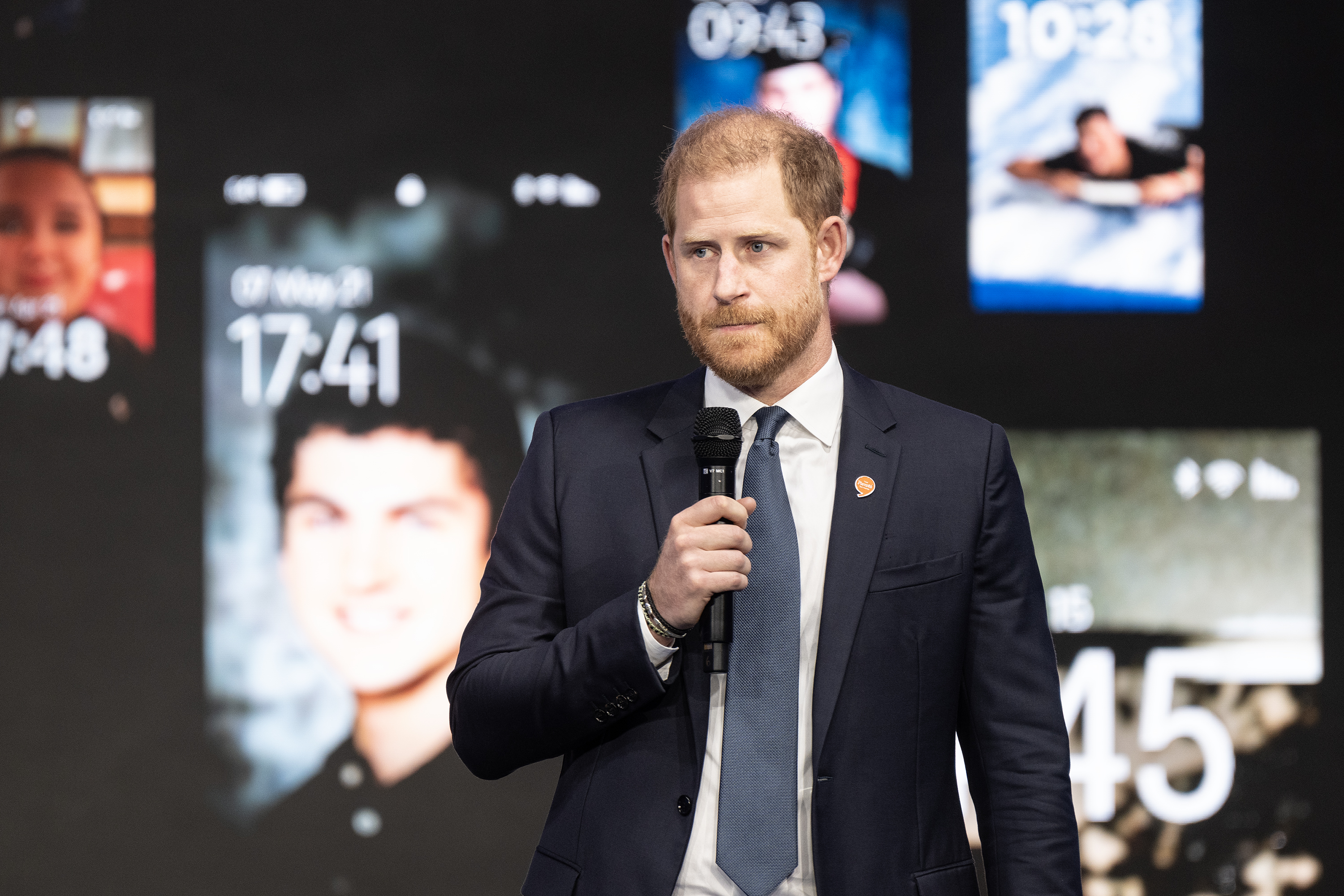 Prince Harry speaking in a mic with photos from phones on a screen behind him