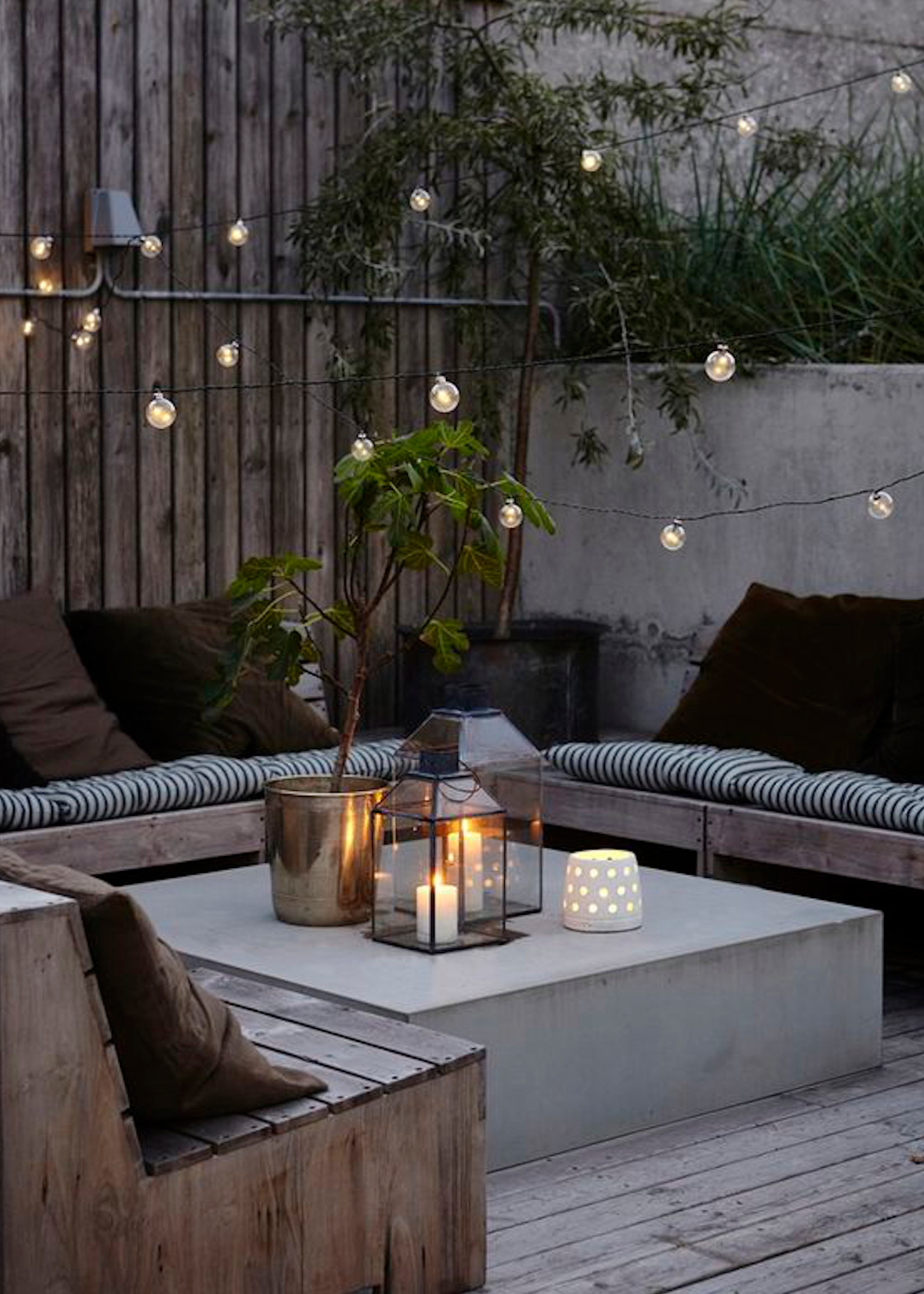 Small patio with low coffee table and festoon lights. On the coffee table are a bunch of candles as well as a large potted plant