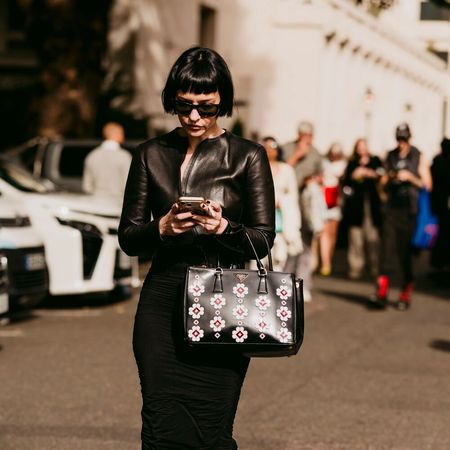 Woman checks phone while wearing an all black outfit. She has a bob, sunglasses, and handbag.