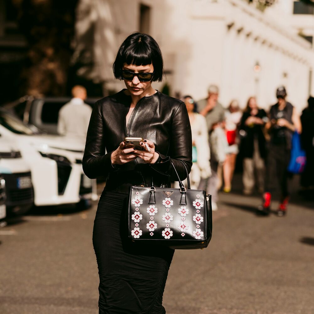 Woman checks phone while wearing an all black outfit. She has a bob, sunglasses, and handbag.