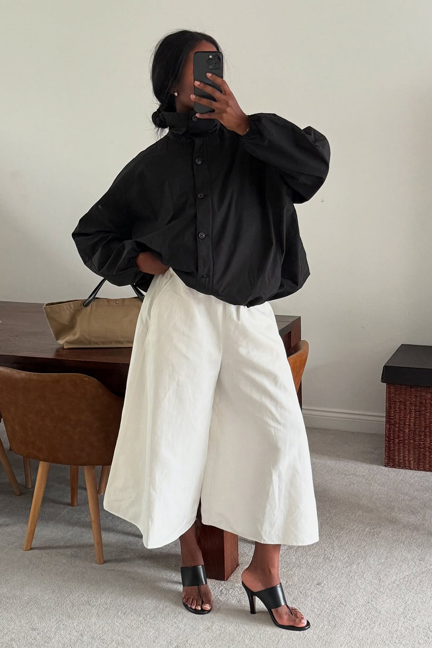 a woman wearing a black funnel neck jacket and white pants and black heels