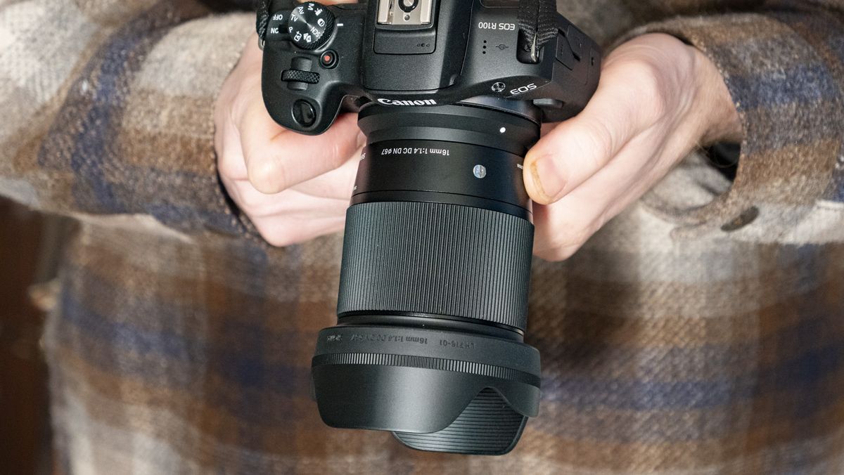 Sigma 16mm F1.4 DC DN Contemporary lens in the hand attached to a Canon EOS R100, checked shirt background