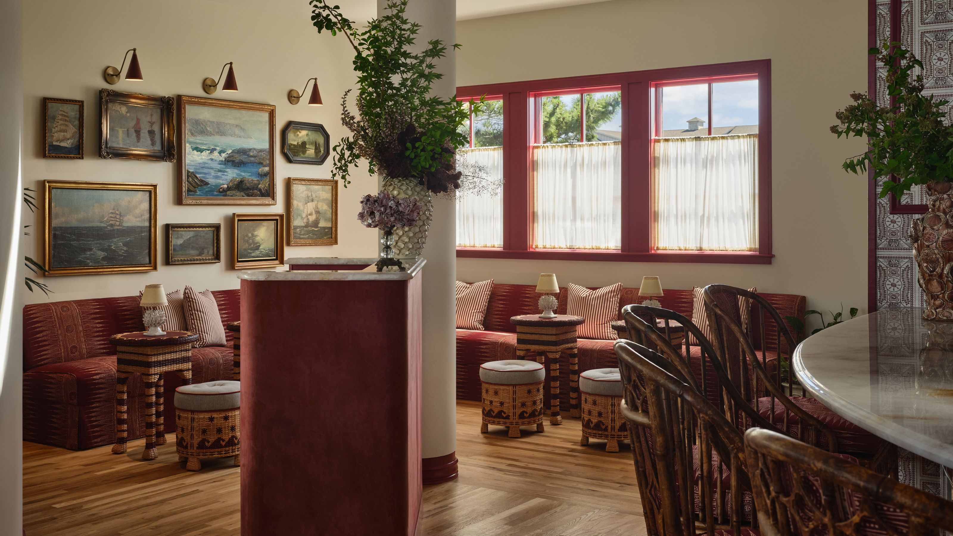 A warm hotel lounge or reception area featuring a gallery wall of framed maritime paintings, patterned red banquette seating, woven stools, and bright red-trimmed windows.