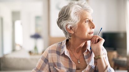 An older woman looks off to the side, thinking, while sitting at her kitchen table.