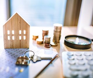 table with wooden model of house, keys, paperwork, piles of coins, calculator and magnifying glass