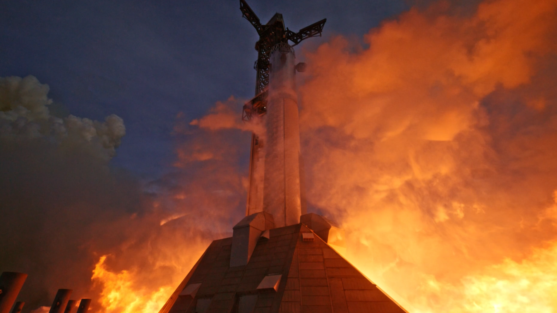 fire erupts from below a giant chrome cylinder with three grid fins, next to a tower.