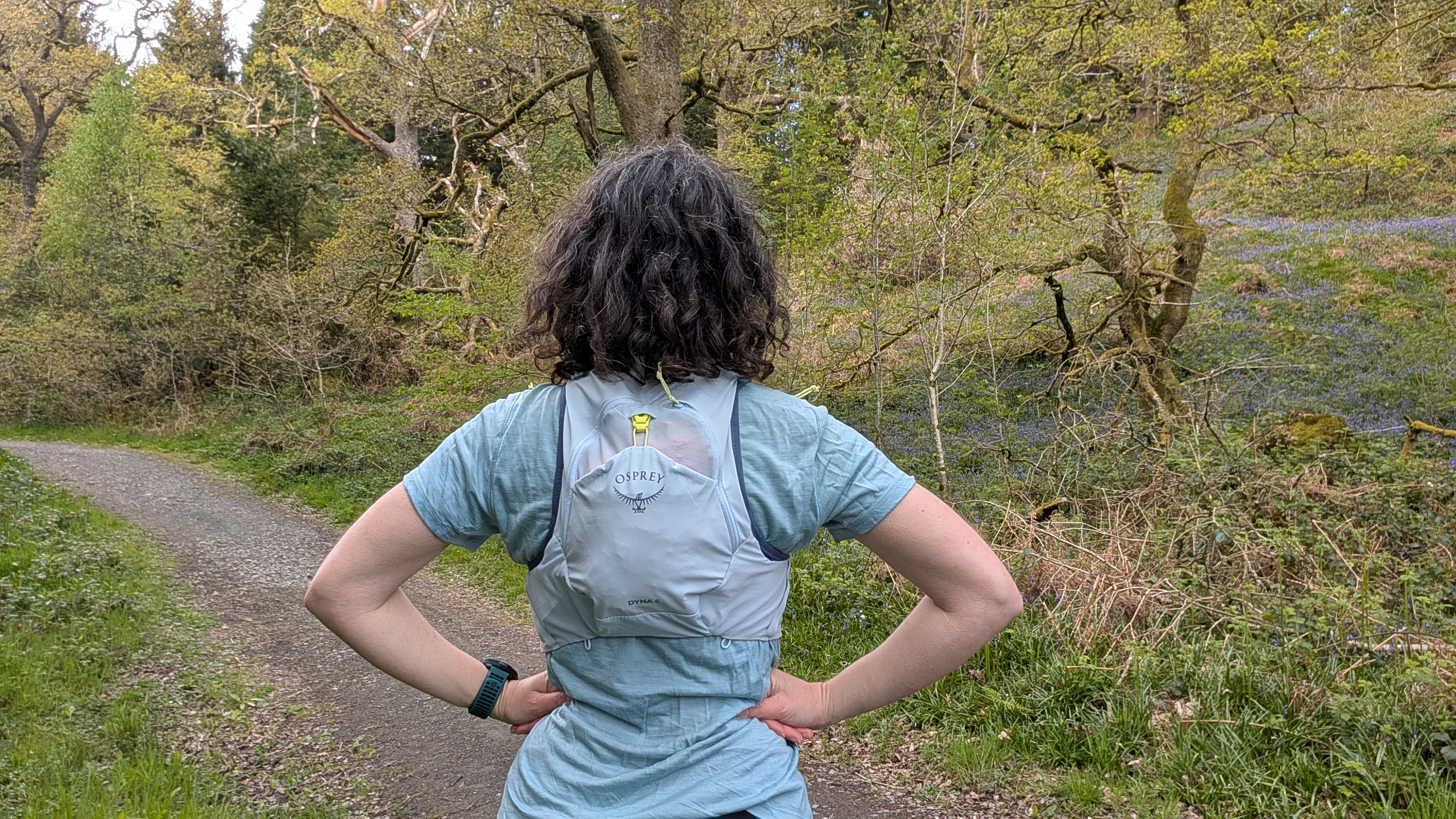 A woman trail runner from behind wearing a light blue T-shirt and Osprey Dyna running vest