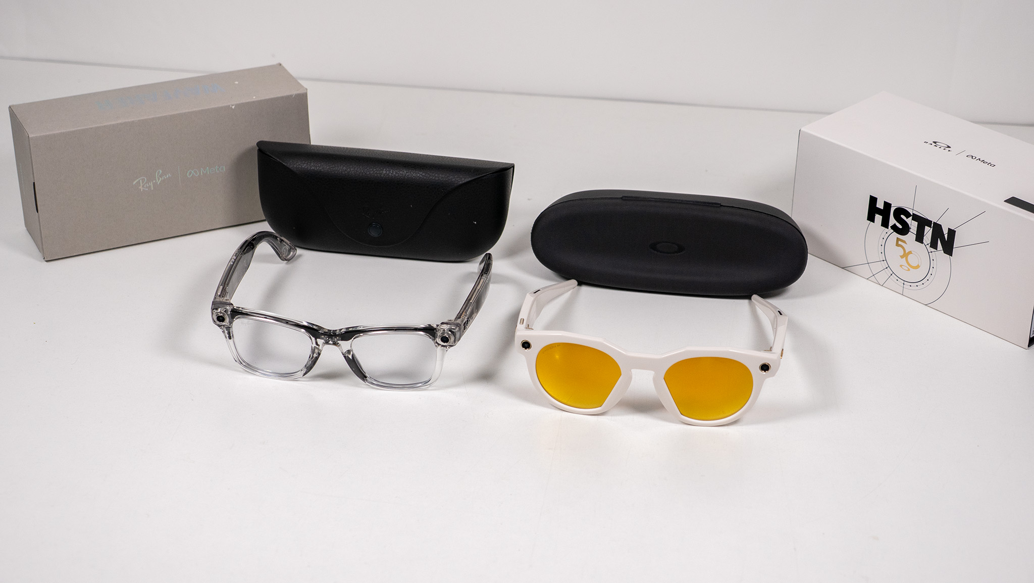 Transparent Ray-Ban Meta Smart Glasses and warm white Oakley Meta HSTN smart glasses with their respective boxes and charging cases on a white table
