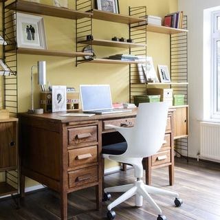 a yellow office study room with wooden floorboards, a wooden desk, white chair on castors and a bespoke wooden and black wire shelving unit